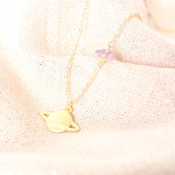 matte gold Saturn pendant on a dainty gold chain with a small amethyst piece on it. the necklace is laying on sparkly pink scarf