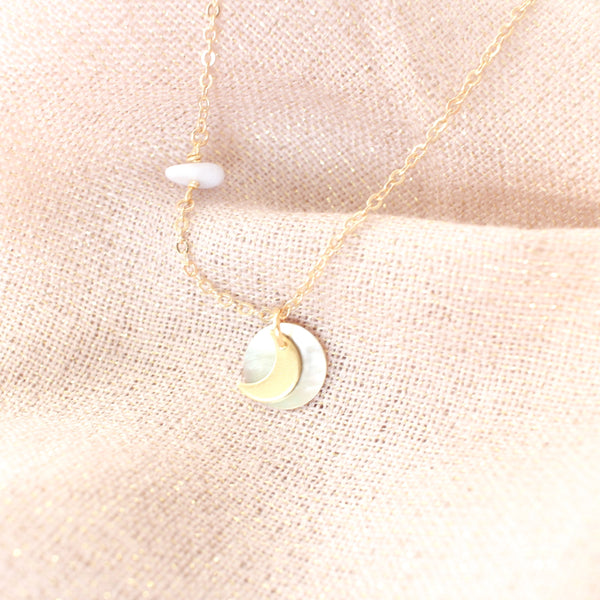 dainty gold necklace with a little moon pendant layered on top of a holographic looking shell pendant, necklace is draped over a sparkly pink fabric.