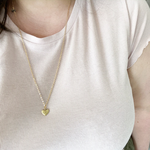 Matte gold heart pendant with an eye carved into it; the pendant is on a dainty gold chain with a piece of tourmalinated quartz on it. the necklace is modelled on a white brunette woman with a light pink shirt