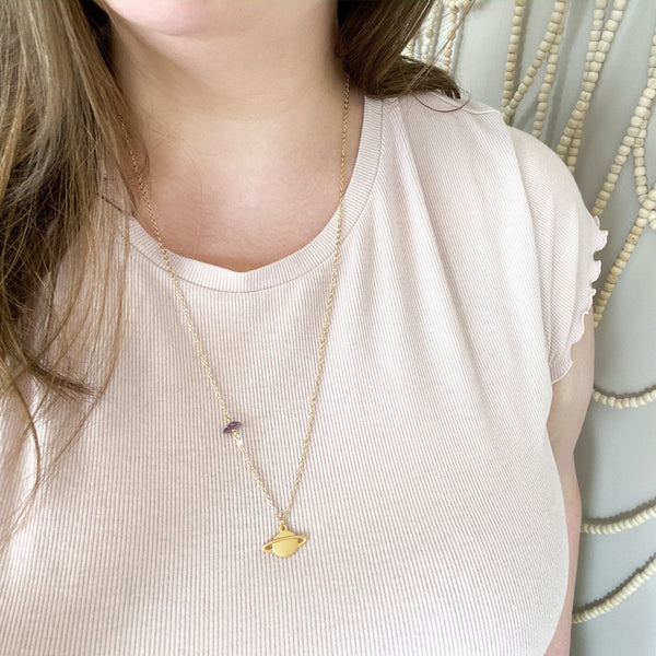 matte gold Saturn pendant on a dainty gold chain with a small amethyst piece on it. the necklace is shown worn by a white brunette woman in a light pink tshirt