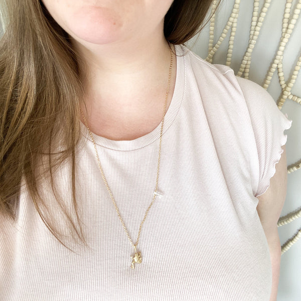 matte gold frog pendant on a dainty gold chain with a little piece of quartz on it. necklace is being worn to show that you can wear the quartz in the front or you can hide it in the back.  the necklace is being modeled by a brunette woman wearing a light pink shirt. in the background is a beaded wall hanging. 