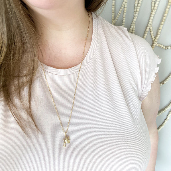 matte gold frog pendant on a dainty gold chain with a little piece of quartz on it. necklace is being worn to show that you can wear the quartz in the front or you can hide it in the back. the necklace is being modeled by a brunette woman wearing a light pink shirt. in the background is a beaded wall hanging.