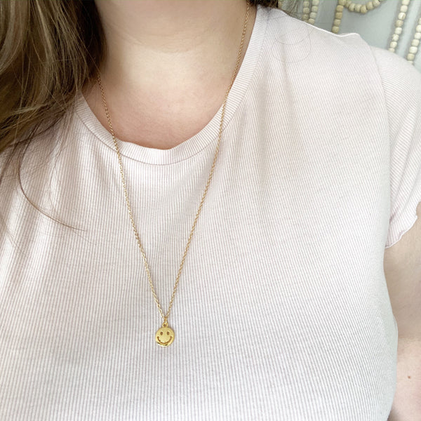 gold smiley face pendant on a dainty gold chain with a little piece of citrine on the chain. necklace is modelled on a caucasian woman with brown hair