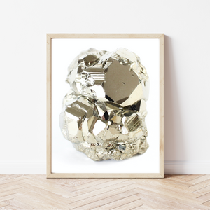 framed photo of a very shiny, metallic pyrite specimen (steely gold colour), there is a hand lettered font underneath that reads "pyrite". the font is the same colour as the crystal. The picture frame is a light natural wood, and the frame is leaning on a white wall, sitting on a light coloured herringbone wood floor.