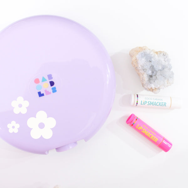 white background flatly image of a purple caboodles case, crystals and lip smackers lip balm. on the caboodles case there is a group of 3 5 petal flower, solid in color with the centre of the flower missing, in various sizes
