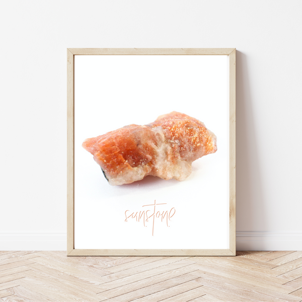 framed photo of a sunstone specimen (earthy orange colour with sparkly parts), there is a hand lettered font underneath that reads "sunstone". the font is the same colour as the crystal. The picture frame is a light natural wood, and the frame is leaning on a white wall, sitting on a light coloured herringbone wood floor.