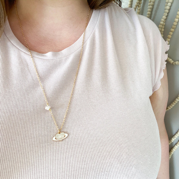 dainty gold chain necklace with a piece of rose quartz on the chain, there is an enamel pendant, a light pink heart with a white ring like a planet. shown worn on a white woman with a light pink t-shirt 