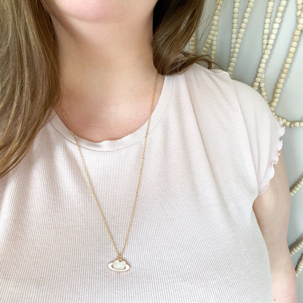 dainty gold chain necklace with a piece of rose quartz on the chain, there is an enamel pendant, a light pink heart with a white ring like a planet. shown worn on a white woman with a light pink t-shirt