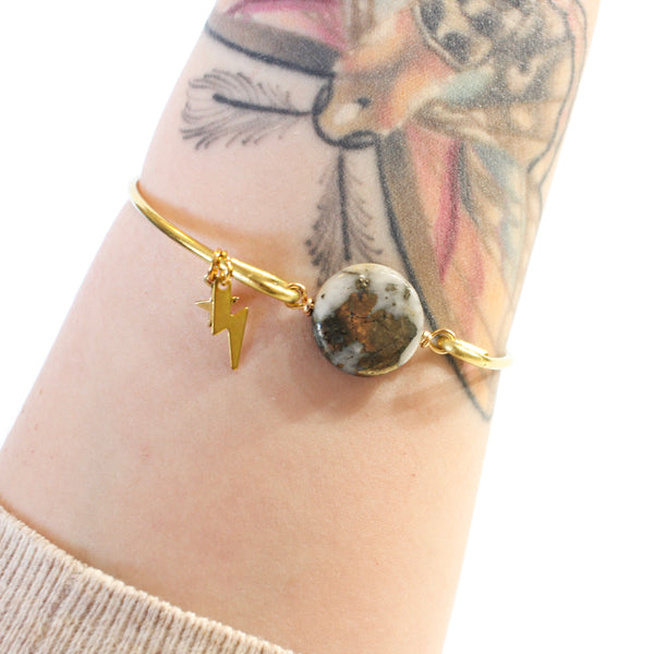 gold wire bangle with two sides held together with a pyrite and quartz coin shaped bead. there are little gold charms on the bangle, a star and a lightning bolt. the pyrite on the bead resembles the marbling on a planet. shown worn on a tattooed wrist, white background.