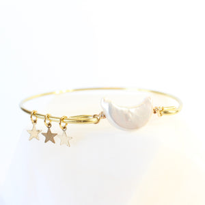 gold bangle with a moon shaped freshwater pearl in the middle.  there are three little gold star charms on the bangle. white background, front 45 degree angle view