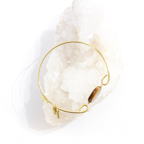 gold coloured bangle with tigers eye bead connecting the two sides. there are three little gold lightning bolt charms on the bangle. top view of bracelet, resting on a bed of a white crystal cluster.  white background