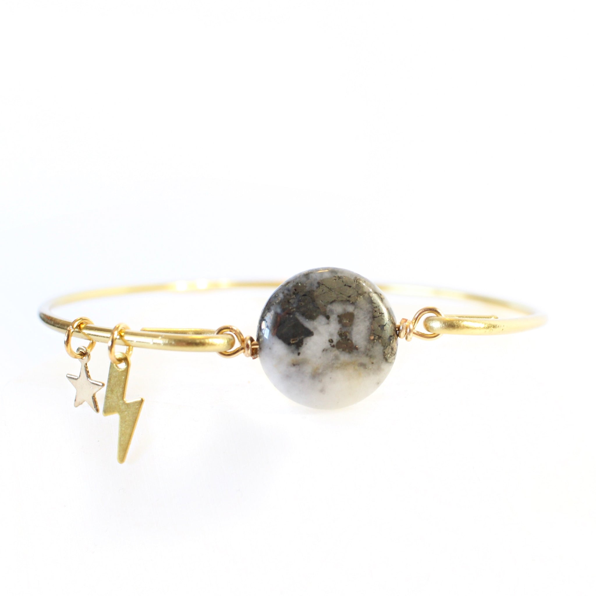 gold wire bangle with two sides held together with a pyrite and quartz coin shaped bead. there are little gold charms on the bangle, a star and a lightning bolt. the pyrite on the bead resembles the marbling on a planet. front view, white background.