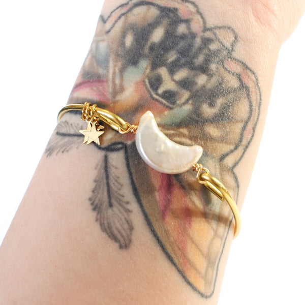 gold bangle with a moon shaped freshwater pearl in the middle. there are three little gold star charms on the bangle. white background, shown worn on a tattooed wrist