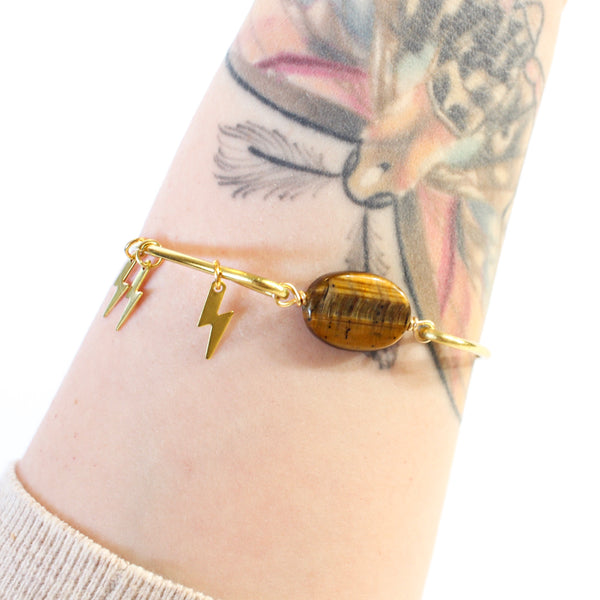 gold coloured bangle with tigers eye bead connecting the two sides. there are three little gold lightning bolt charms on the bangle. pictured worn on a tattooed wrist, white background