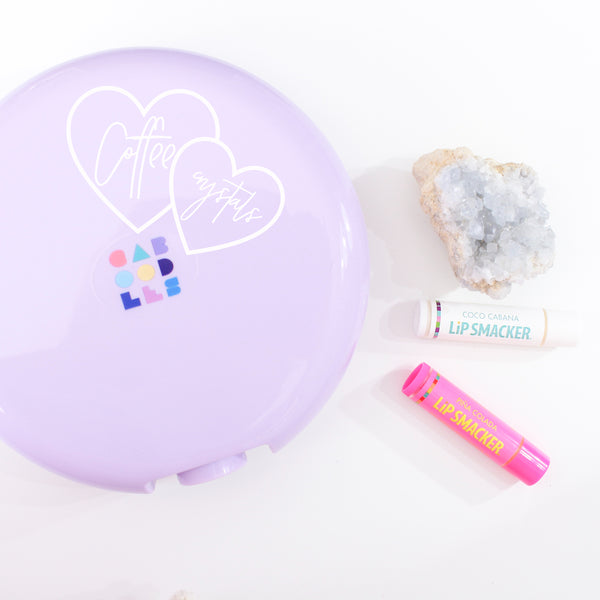 round purple caboodles case on a white background, on the case there is a white decal with two interlocking hearts, one says coffee, and other says crystals. beside the case there is a soft blue crystal cluster, and two  Lip Smackers lip balm tubes