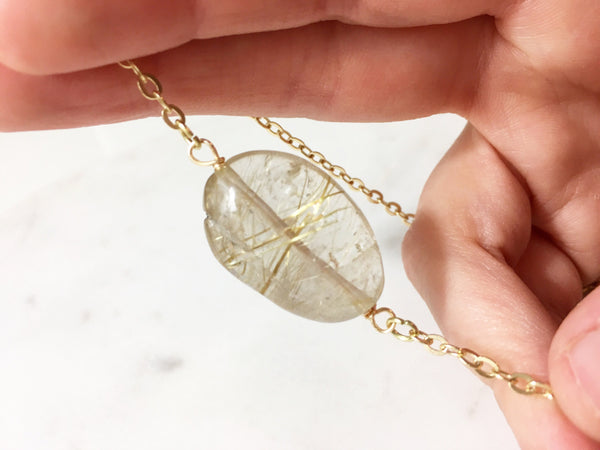 caucasian woman's hand holding thin gold chain with a gold rutilated quartz oval pendant, marble background