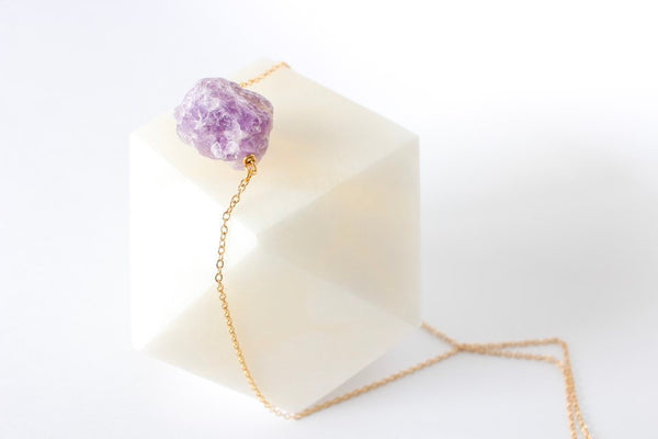 deep purple raw amethyst chunk on a thin gold chain placed on geometric marble piece. white background, side view