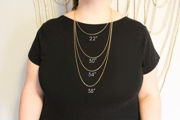 caucasian plus sized woman wearing black shirt modelling different lengths of thin gold chain. neutral background with beaded wall hanging. 22", 30", 34", and 38" shown