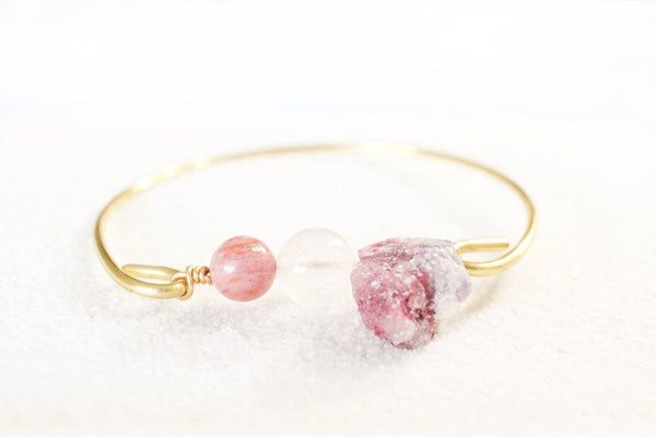 front view of gold brass self love bangle, with rhodonite, rose quartz, and raw pink tourmaline laying on white sand