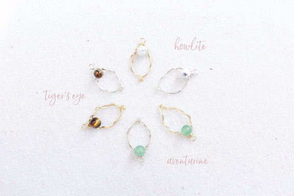 6 wire yoni charms in silver and gold with different gemstone clitorises of howlite, tigers eye and aventurine. in a circle laying on white sand