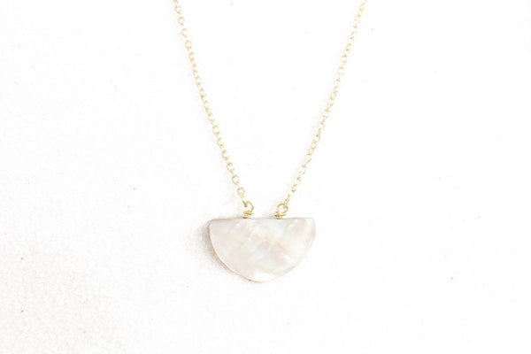 silvery iridescent half moon shaped seashell pendant on a thin gold chain, top view. laying on white sand