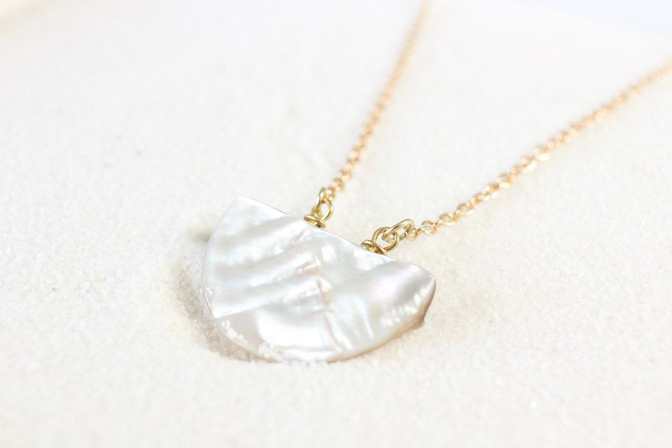 45 degree angle view of a silver iridescent half moon shaped seashell pendant on a thin gold chain, laying on white sand