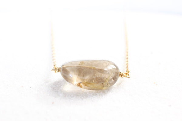 bottom view of gold rutilated quartz necklace on white sand, with light shining through the quartz