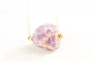 straight-on view of purple raw amethyst chunk on a dainty gold chain, laying on white sand