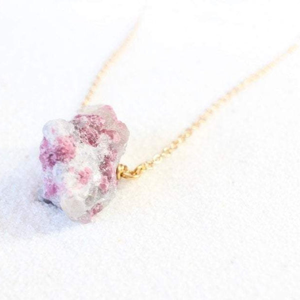 45 degree angle view of a large raw pink tourmaline chunk on a dainty gold chain, laying on white sand