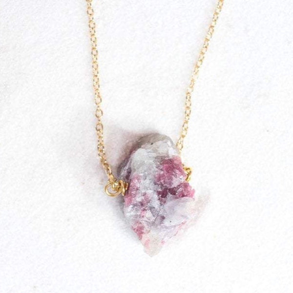 top view of a large raw pink tourmaline chunk on a thin gold chain, laying on white sand