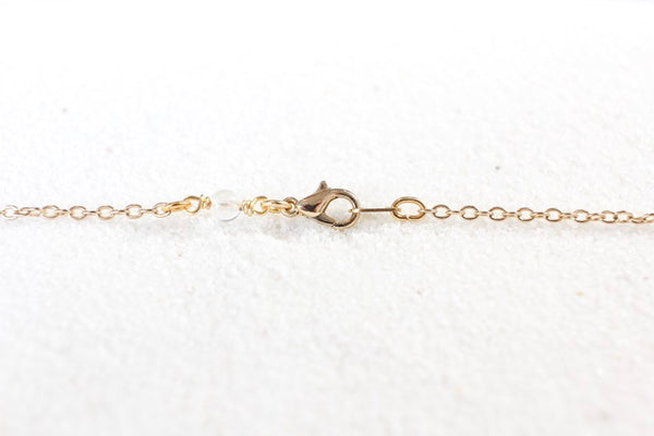 dainty gold chain laid across white sand. shows clasp and small quartz bead near clasp