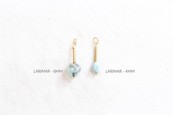 2 larimar pendants of different sizes with gold detail, laying on white sand