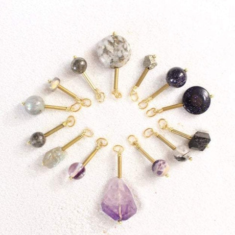 pyrite, blue goldstone, black tourmaline, tourmalinated quartz, amethyst, fluorite and labradorite pendants with gold brass detail, laying on white sand in a starburst figuration