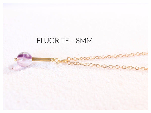 fluorite pendant with gold brass detail, strung on a dainty gold chain and laying on white sand