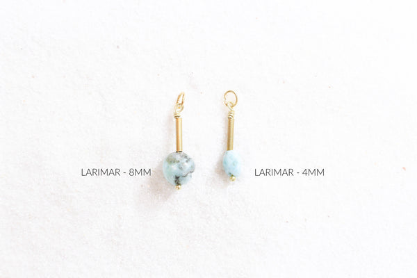 2 sizes of larimar pendants 8mm and 5mm with gold detail, laying on white sand