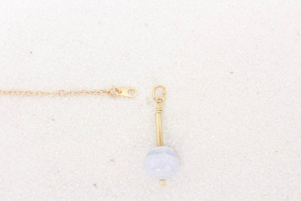 clasp tab attached to thin gold necklace is shown laying on white sand, with a healing charm beside it. photo shows how charms fit onto the chain