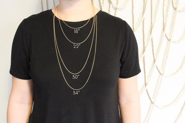 average sized caucasian woman in black shirt modelling different length gold chains. background is neutral with beaded wall hanging. 18", 22", 30", and 34" shown