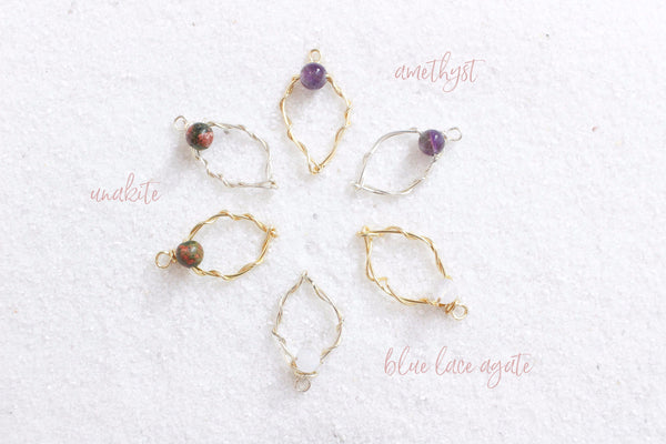 6 wire yoni charms in silver and gold with different gemstone clitorises of amethyst, blue lace agate, and unakite. in a circle laying on white sand