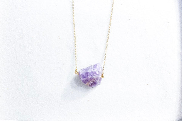 large raw deep purple amethyst chunk on a thin gold chain, top view laying on white sand