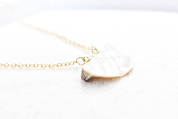 side view: a silvery iridescent half moon shaped seashell pendant on a dainty gold chain laying on white sand