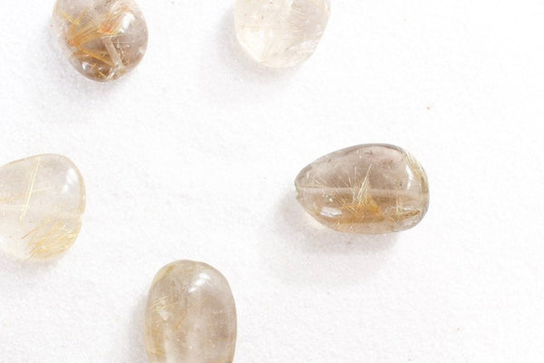 5 pieces of gold rutilated quartz laid in a circle on white sand, showing different shapes and colours (clear to a smokey colour)