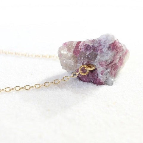 side view of a large raw pink tourmaline chunk on a thin gold chain, laying on white sand