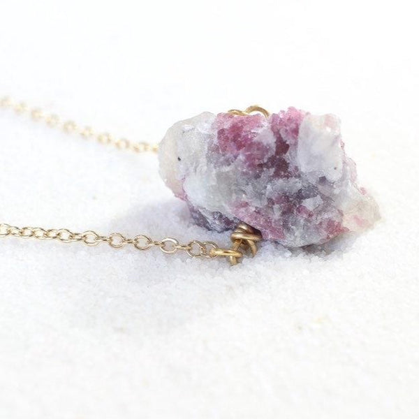 right side view of a large raw pink tourmaline chunk on a thin gold chain laying on white sand