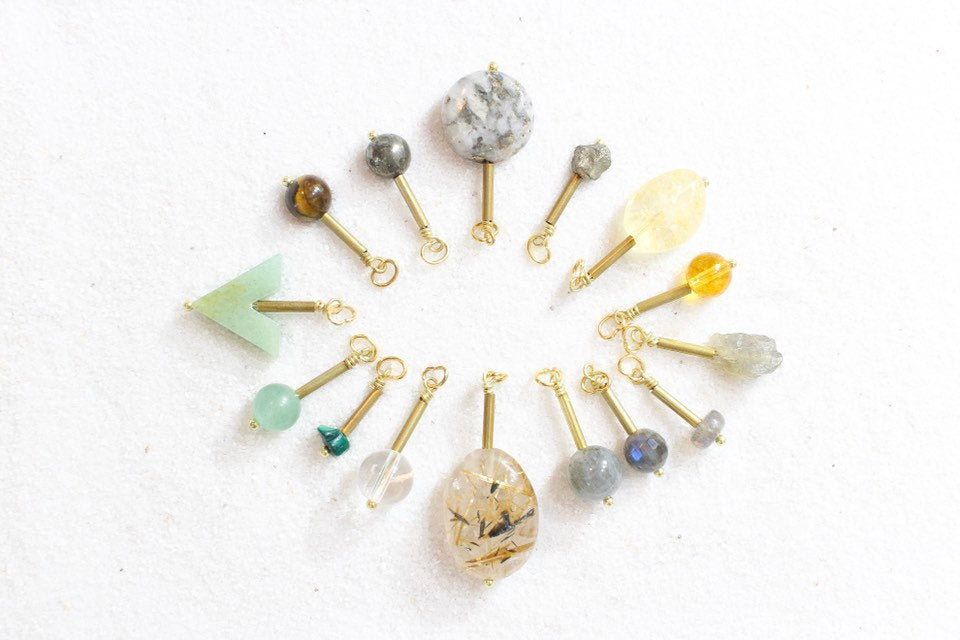 pyrite, citrine, labradorite, gold rutile quartz, clear quartz, malachite, aventurine, and tigers eye pendants with gold brass detail, laid out in a sunburst shape on white sand