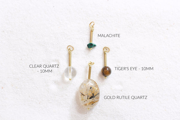 malachite, tigers eye, gold rutile quartz, and clear quartz pendants with gold brass detail laid out on white sand