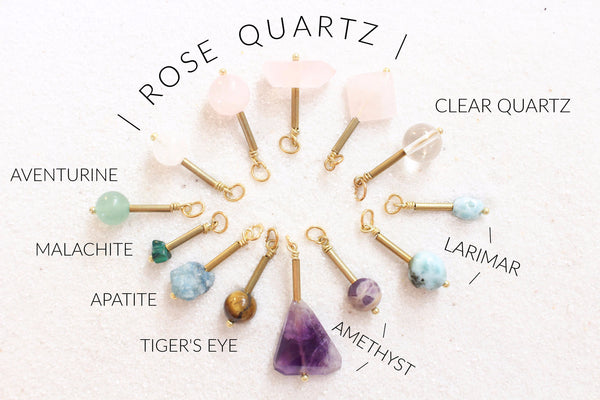 rose quartz, clear quartz, larimar, amethyst, tiger's eye, apatite, malachite, aventurine pendants all with gold detail, laying in a sunburst shape on white sand. crystals are labelled accordingly and are grouped together