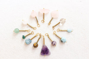 rose quartz, clear quartz, larimar, amethyst, tiger's eye, apatite, malachite, aventurine pendants, all with gold detail, laying in a sunburst shape, on white sand