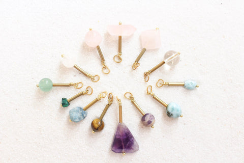 rose quartz, clear quartz, larimar, amethyst, tiger's eye, apatite, malachite, aventurine pendants, all with gold detail, laying in a sunburst shape, on white sand